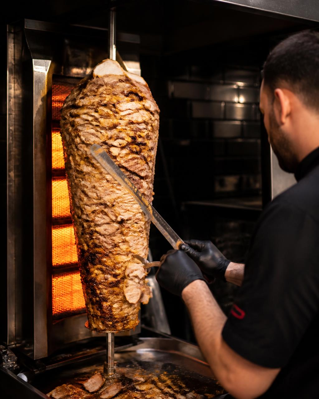 Chef carving thin slices of pork gyros from a vertical rotisserie spit